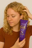 Blonde woman with natural curly hair applying Aussie Miracle Curls Frizz Taming Cream for defined, frizz-free curls – enriched with coconut and Australian jojoba oil, perfect for curly hair care in South Africa.