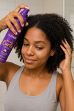 A young woman with natural curls applying Aussie Miracle Curls Mousse with coconut and jojoba oil to her hair in a bathroom for defined, frizz-free curls.