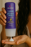 A person with damp hair dispensing Aussie 3 Minute Miracle Moist Deep Conditioner into their hand, holding the purple bottle upside down.